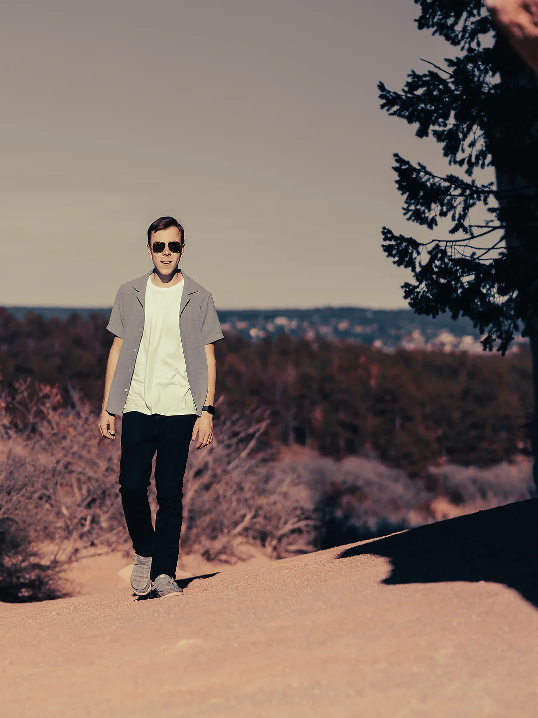 Young man in an open grey shirt and aviator sunglasses walking toward the camera along a sandy ridge, a wooded valley and distant town behind