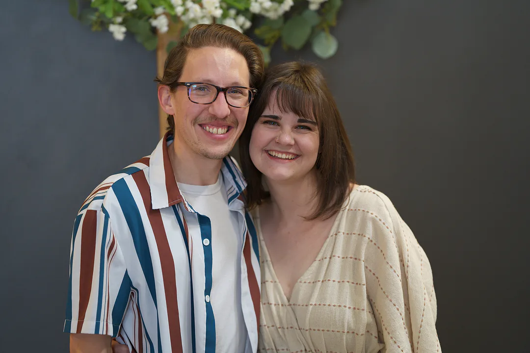 A couple smiling cheek to cheek against a grey wall, he in a striped shirt and glasses, she in a cream knit top, white flowers above them