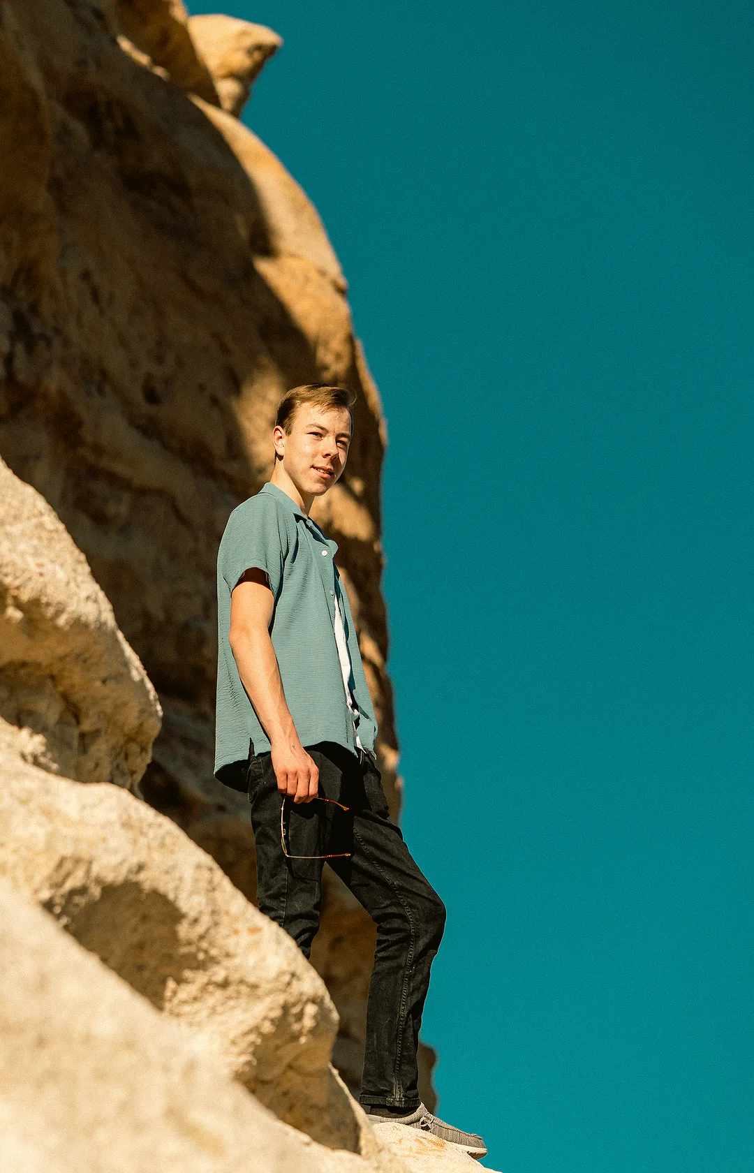 Young man standing on a sandstone ledge against a deep blue sky, sunglasses in one hand, turning to look back at the camera