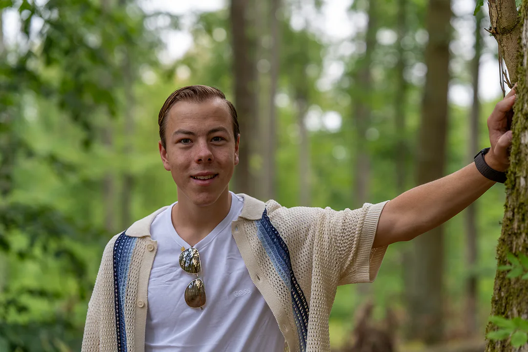 Young man leaning one arm against a tree in a green forest, smiling in a white tee and crochet cardigan