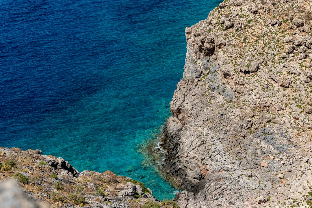 Rocky cliff seen from above, dropping into turquoise water that deepens to dark blue