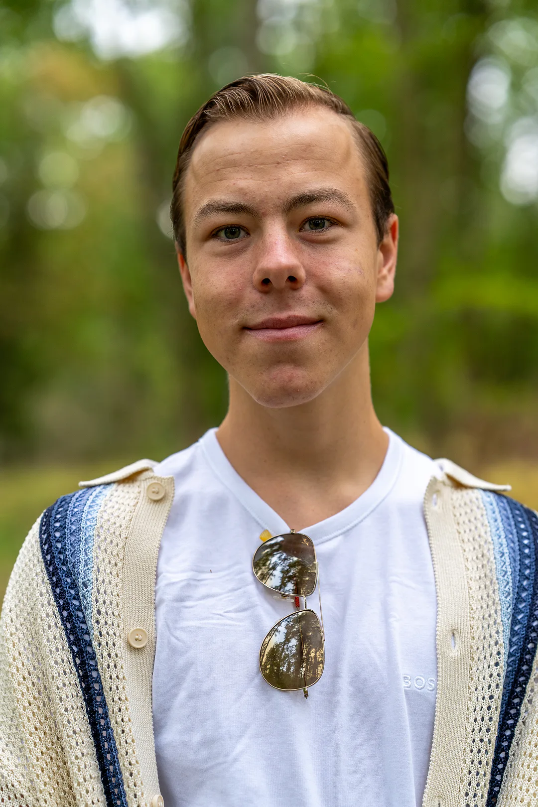 Head-and-shoulders portrait of a young man in a cream knitted cardigan, aviator sunglasses hanging from the neck of his white tee, green trees blurred behind him