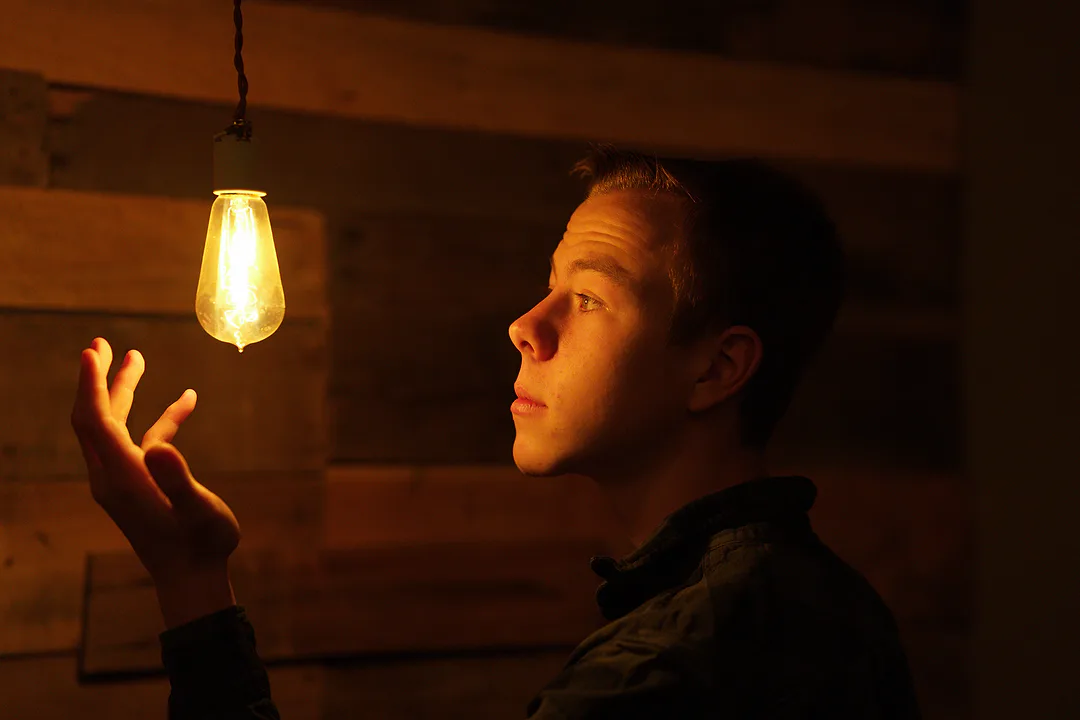 Young man in profile lit only by a hanging Edison bulb, one hand raised toward it against dark barn wood