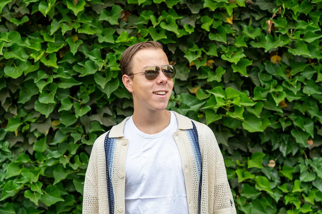Young man in aviator sunglasses laughing and looking off to the side, standing before an ivy-covered wall
