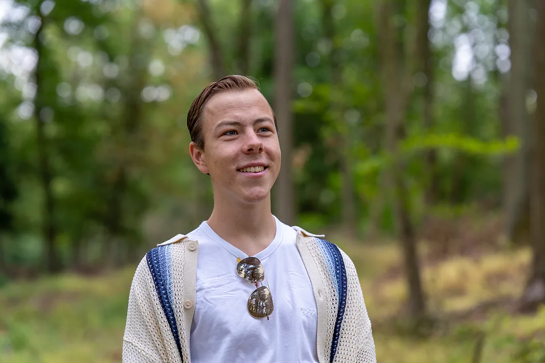Young man in a cream and blue knitted cardigan over a white tee, sunglasses hooked at his collar, smiling and looking off to one side in a sunlit wood