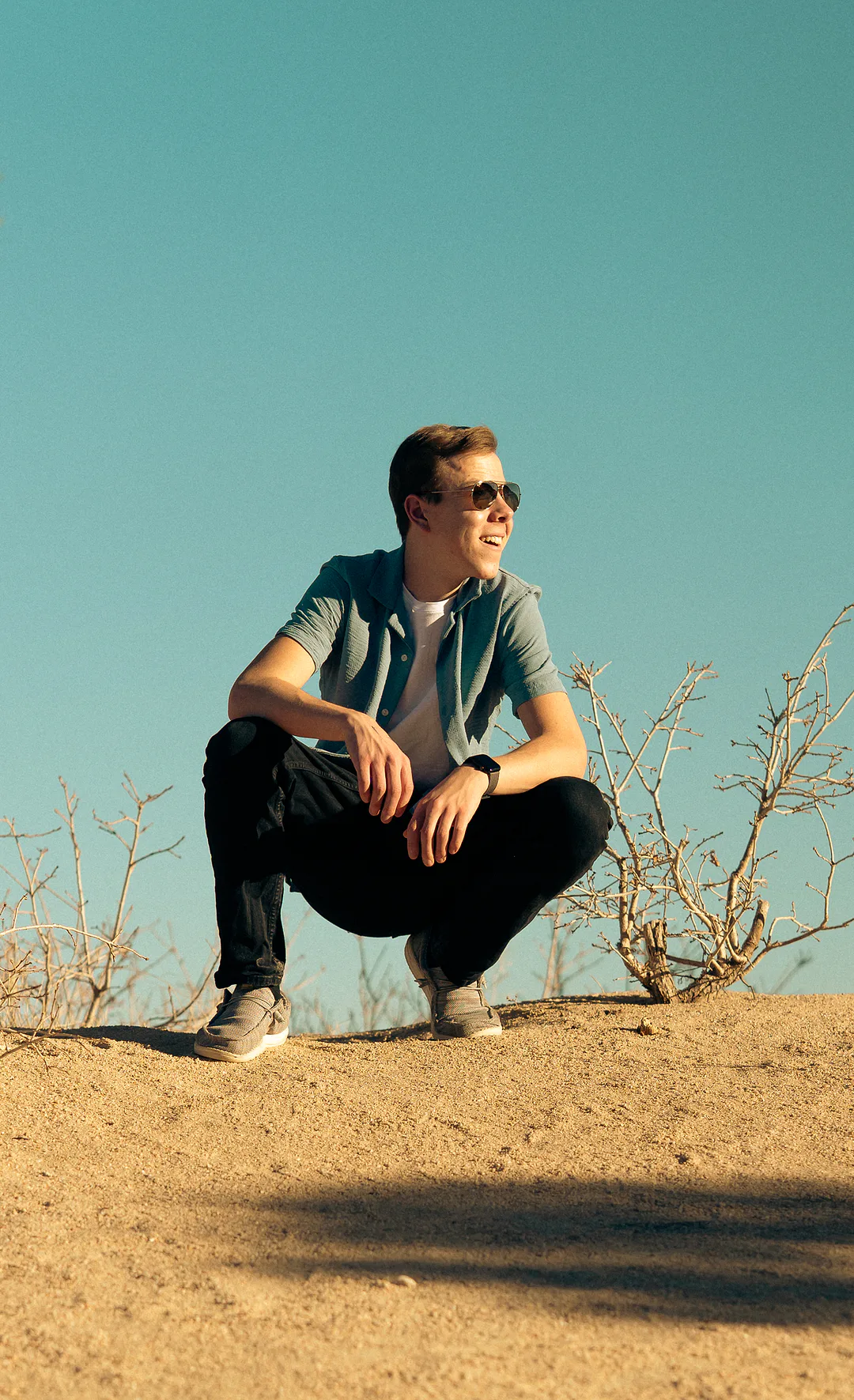 Young man crouching on a sandy rise against a clear turquoise sky, smiling off to one side in sunglasses