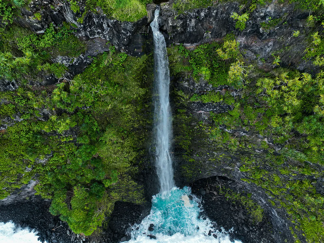 Aerial view of a waterfall dropping down a green cliff into a turquoise pool where it meets the sea