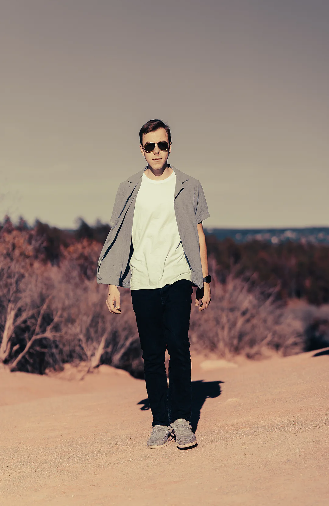Full-length portrait of a young man in a grey overshirt and aviator sunglasses standing on open sand among bare winter brush