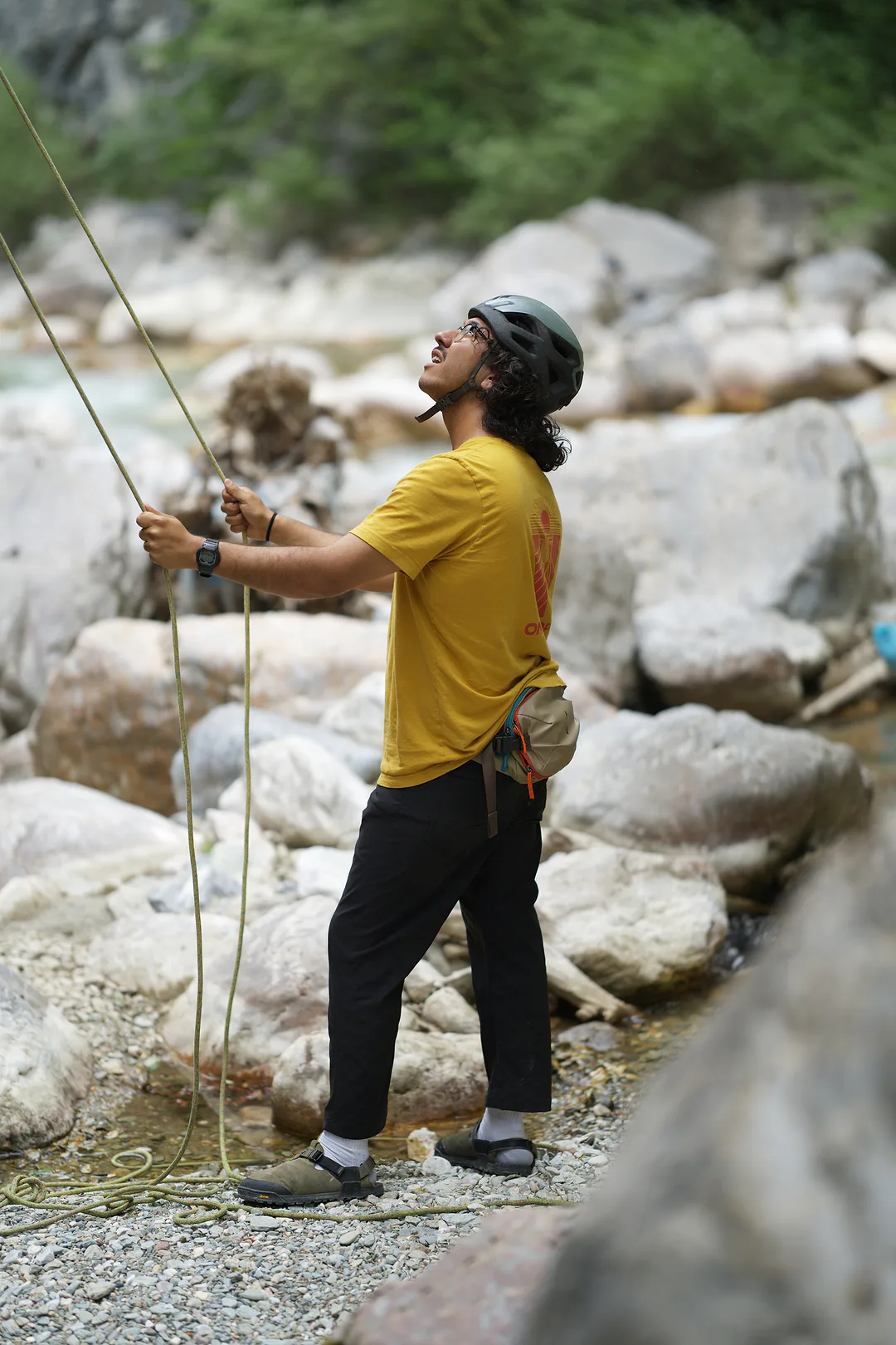 Man in a yellow shirt and climbing helmet feeding rope through his hands and looking up, standing on pale boulders beside a canyon river