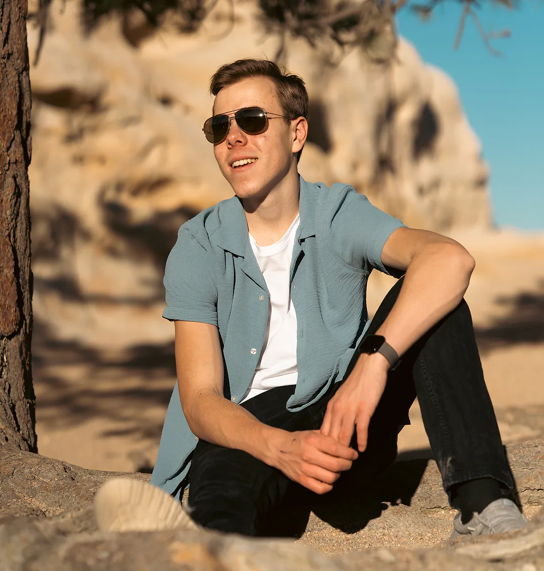 Young man in a teal shirt sitting on rock in dappled shade, laughing, a pale sandstone bluff and blue sky behind him