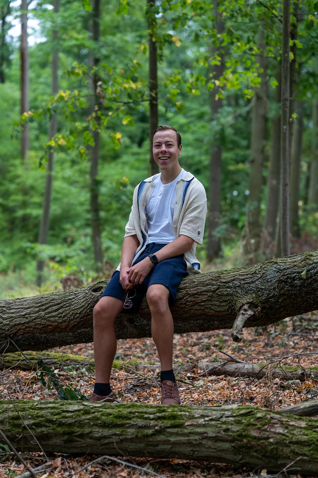 Young man sitting on a fallen log in the woods, hands together, smiling toward the camera