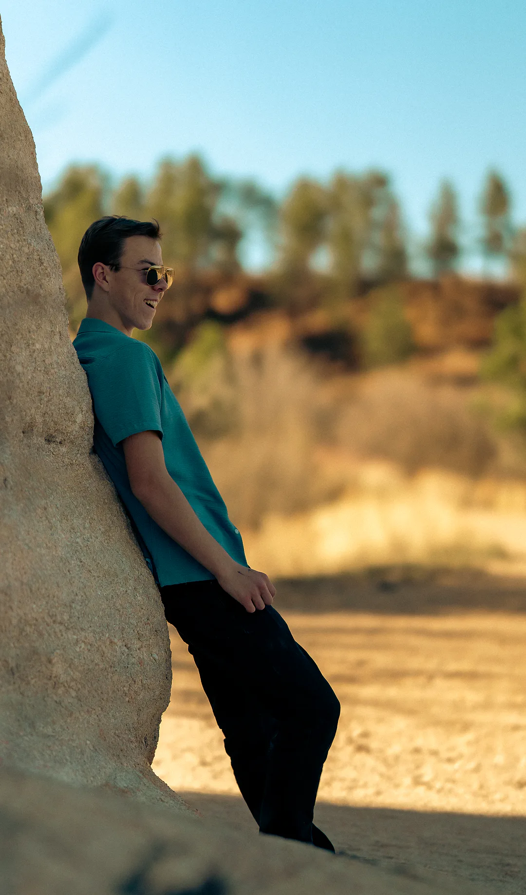 Young man in a teal shirt leaning back against a sandstone boulder, laughing, a sunlit golden field stretching behind him
