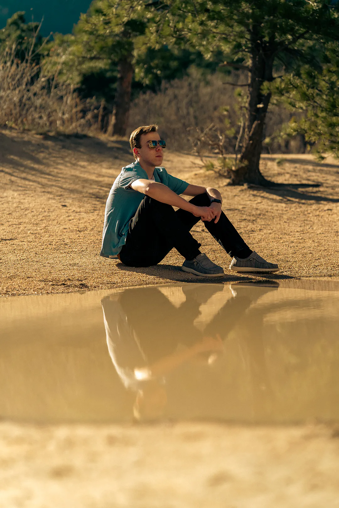 Young man sitting on packed sand at the edge of a still pool, his reflection in the water and pines behind him in low golden light