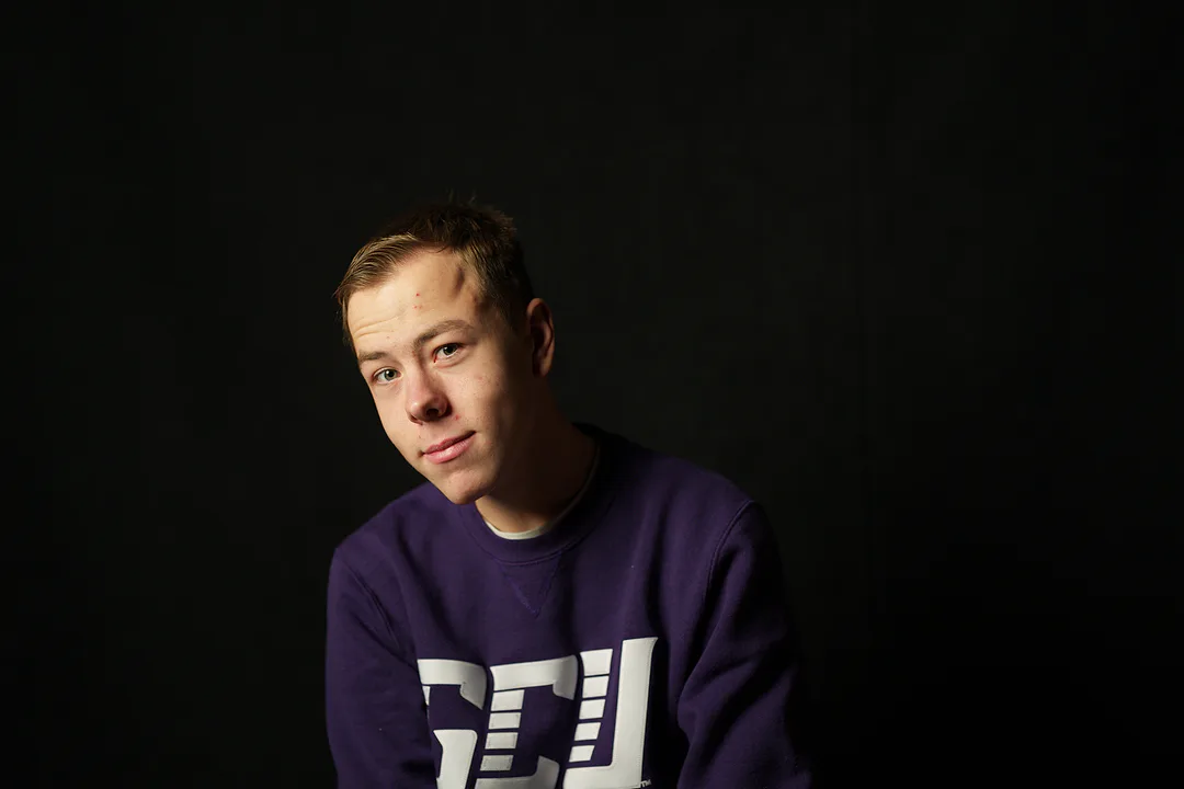 Studio portrait of a young man in a purple university sweatshirt against a black background, half his face in soft light