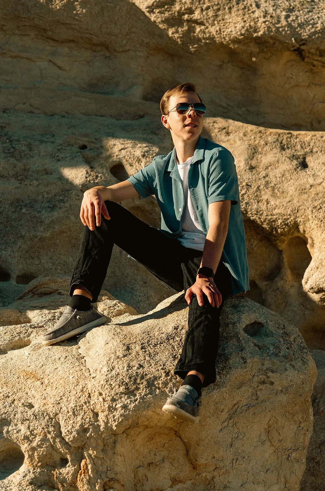 Young man in a teal camp shirt and sunglasses sitting on warm sandstone, one arm resting across his knee in late afternoon light