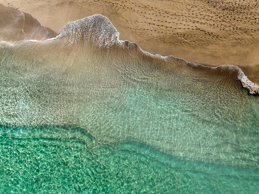 Aerial view of clear turquoise shallows meeting a tan beach, a line of white foam and footprints across the wet sand
