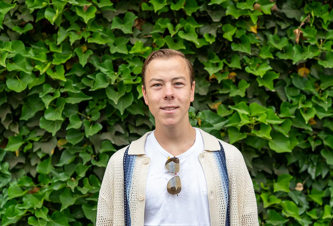 Young man in a cream crochet cardigan over a white tee, sunglasses hooked at his collar, smiling in front of a wall of ivy
