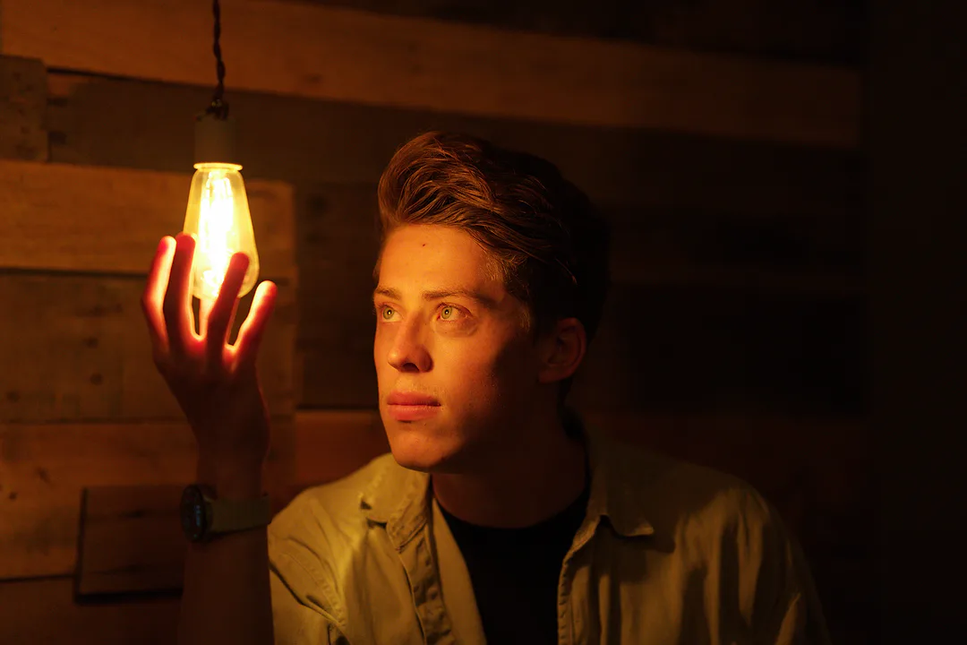 Young man in a dim wood-panelled room holding his hand beside a hanging Edison bulb, its warm light glowing through his fingers