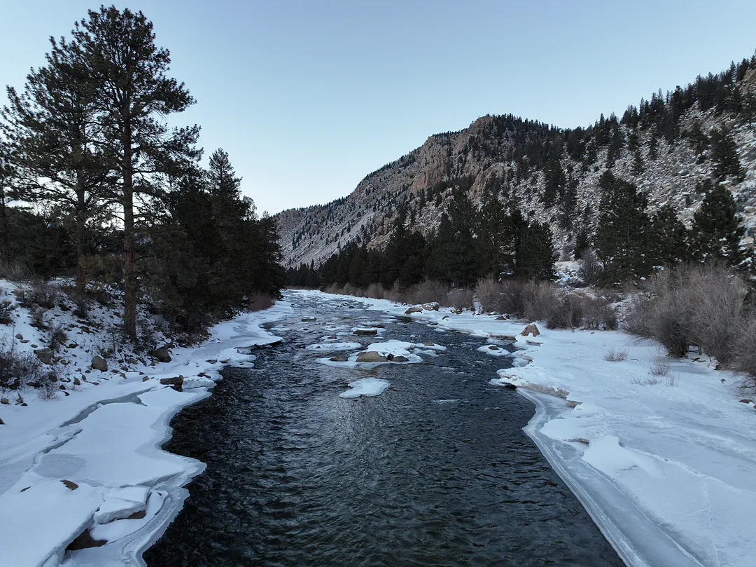 A dark river running between snow-covered banks in a pine valley, a rocky ridge catching the last of the light