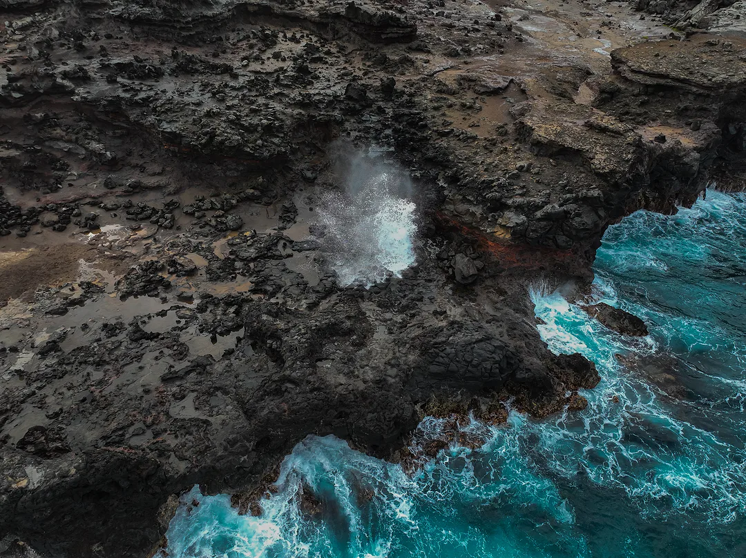 Aerial view of a black volcanic shoreline where surf bursts white through a blowhole and turquoise water breaks along the rock